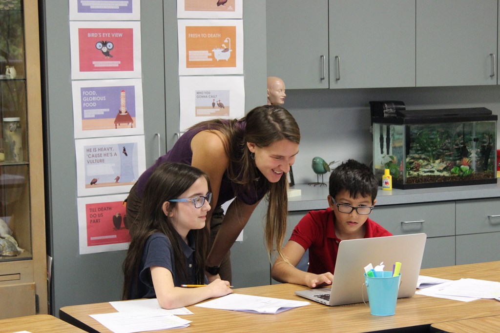 Teacher working with students in the classroom using a laptop and activity handouts