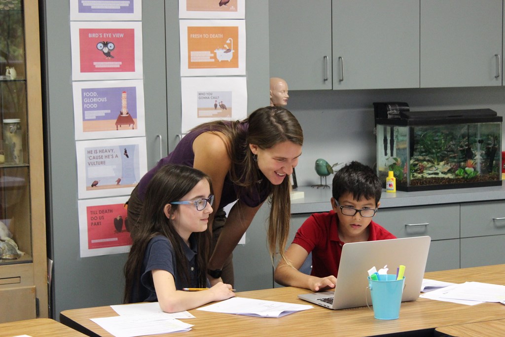 Teacher working with students in the classroom using a laptop and activity handouts